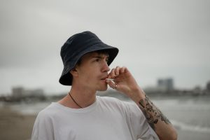 Joven con sombrero y tatuajes fuma en una playa