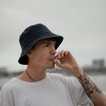 Joven con sombrero y tatuajes fuma en una playa